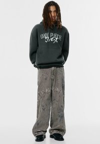 Sötétzöld kapucnis pulóver, rajta a "ONE PIECE" felirattal, szürke, túlnőtt, festékspricceléses nadrággal és fekete csizmával párosítva, laza szabással.