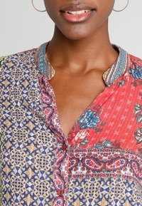 Multicolored blouse with floral and geometric patterns, featuring a button placket and a textured, embroidered collar. Cotton fabric.