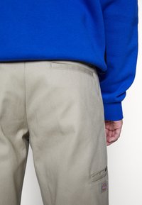 Pantalones beige con una textura suave, que cuentan con un bolsillo de parche y costuras sutiles. Combinados con un sudadera azul vibrante.