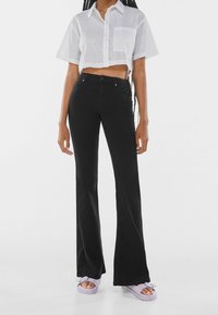 Jeans negros acampanados combinados con una camiseta blanca corta que presenta patrones de ojales. La modelo lleva sandalias deslizantes de color púrpura claro.