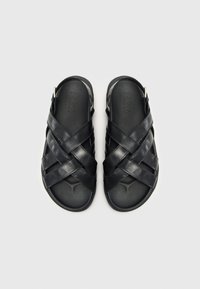 Sandali in pelle nera con cinturini incrociati, design slip-on e una soletta testurizzata. Il nome del marchio "ZIGN" è impresso sulla soletta.