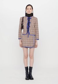 Gekruiste tweed jurk met blauwe accenten, voorzien van een hoge kraag, strikjes en een zak. Model draagt zwarte kniehoge laarzen.