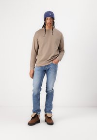 Beige hettegenser, blå jeans og brune støvler med lysere detaljer. Modellen har på seg en blå lue og står foran en hvit bakgrunn.