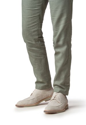 Zapatos de lino gris claro con textura suave, con punta redondeada y tres ojales. Usados con pantalones ajustados de color verde oliva.