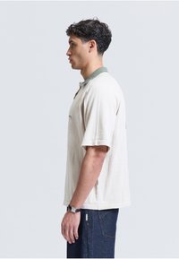Jeune homme debout de profil portant une chemise crème à manches courtes avec un col vert et un jean bleu foncé sur un fond uni.