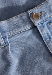 Lichtblauwe denimstof met zichtbare textuur, voorzien van een messing knoop en gele stiksels langs de tailleband en zakken.