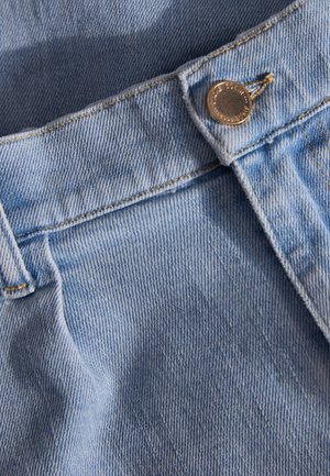 Hellblaues Denimgewebe mit sichtbarer Textur, ausgestattet mit einem Messingknopf und gelben Nähten entlang des Taillenbunds und der Taschen.