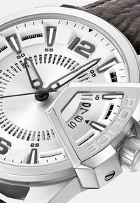 Zilveren roestvrijstalen horloge met een gestructureerd, donkerbruin leren band. Voorzien van opvallende uurmarkeringen, verfijnde wijzerplaatpatronen en details aan de zijkant.