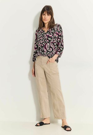 Schwarze Bluse mit langen Ärmeln und pink-weißer Paisley-Musterung, kombiniert mit beigefarbenen weit geschnittenen Hosen und schwarzen Riemensandalen.