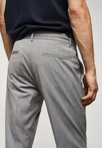 Pantaloni grigi a righe con una vestibilità su misura, dotati di tasche posteriori e una texture liscia. Indossati con una maglia scura che mette in evidenza il punto vita.