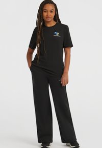 Camiseta negra de manga corta con un logotipo colorido en el pecho izquierdo, combinada con pantalones negros de corte holgado. Textura de tela suave.