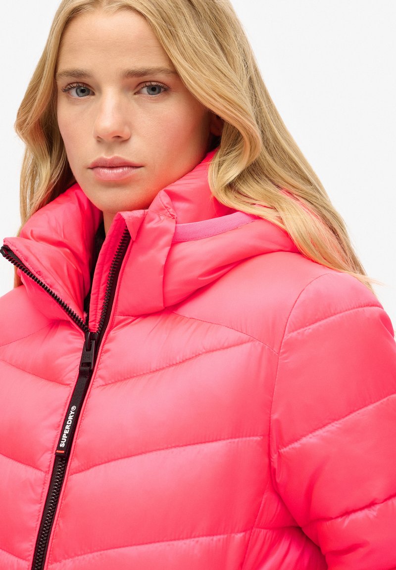 Rosa gepolsterte Jacke mit hohem Kragen und Reißverschluss. Verfügt über gestickte Abschnitte und einen markierten Reißverschluss-Anhänger. Glatte, glänzende Textur.