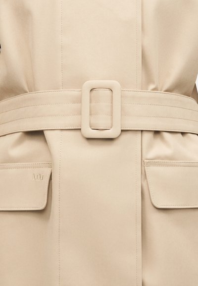 Μπεζ ένδυμα με ζώνη και ορθογώνια αγκράφα, καθώς και τσέπες με καπάκι, μία από τις οποίες φέρει ανάγλυφα τα αρχικά "MM".