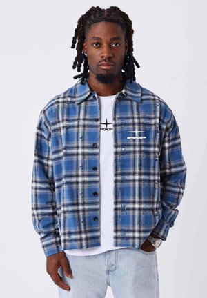 Homme aux cheveux tressés portant une chemise à carreaux bleue boutonnée par-dessus un t-shirt blanc et un jean en denim clair, debout devant un fond blanc uni.