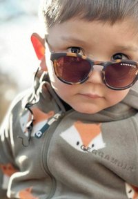 Harmaa fleece-takki, jossa on oransseja kettu-kuvioita; lapsi, jolla on pyöreät aurinkolasit kilpikonnakuosisella kehys. Sileä tekstuuri, valaistu tausta.