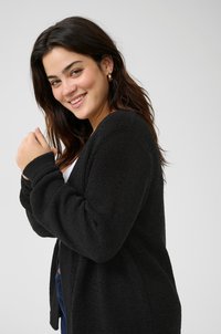 Cardigan noir en tricot avec un motif texturé, à l'avant ouvert et à manches longues, porté sur un haut blanc, sur un fond uni.