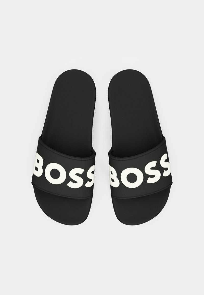 Schwarze Gummisandalen mit breitem Riemen, der den auffälligen weißen Schriftzug "BOSS" trägt. Glatte Textur, abgerundete Form und flaches Sohlendesign.