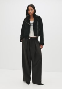 Giacca in shearling nera con un design corto, chiusura con zip e maniche ampie, abbinata a pantaloni scuri a vita alta e tacchi neri a punta.