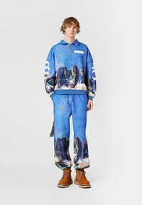 Sudadera azul con cremallera y estampado de paisaje rocoso; pantalones azules a juego; tela texturizada; botas de cordones marrones; corte holgado.