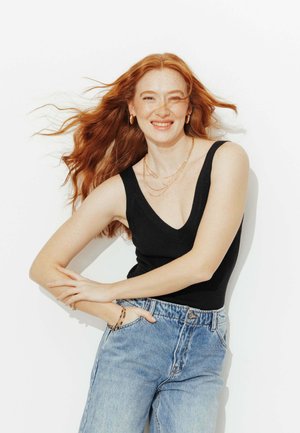 Femme aux cheveux roux portant un top noir sans manches et un jean bleu, souriant avec les bras croisés, cheveux flottant sur un fond blanc.