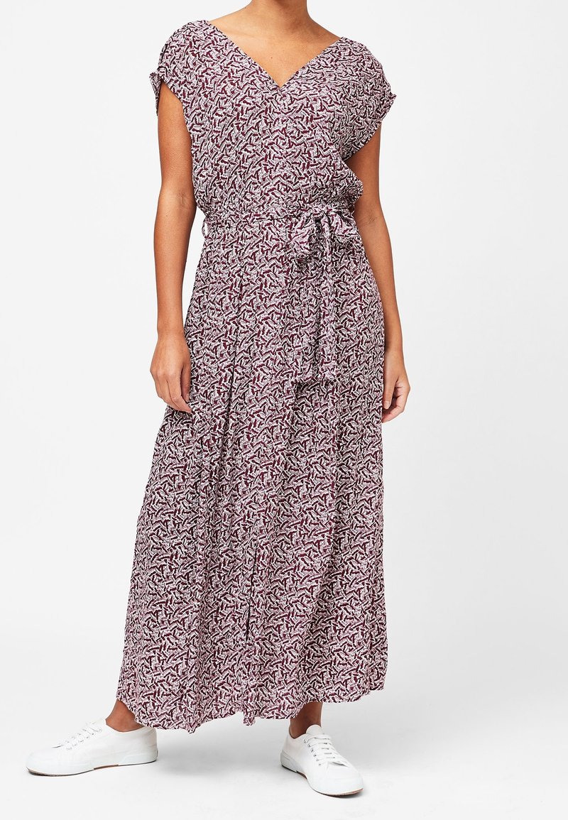 Femme portant une robe maxi sans manches à motifs avec un col en V, une ceinture à la taille et des baskets blanches, debout devant un fond uni.