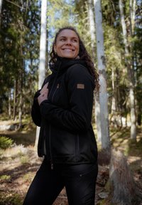Svart softshell-jacka med hög krage, dragkedja fram och fickor på sidorna. Har strukturerad tyg och en märkeslogotyp på bröstet.