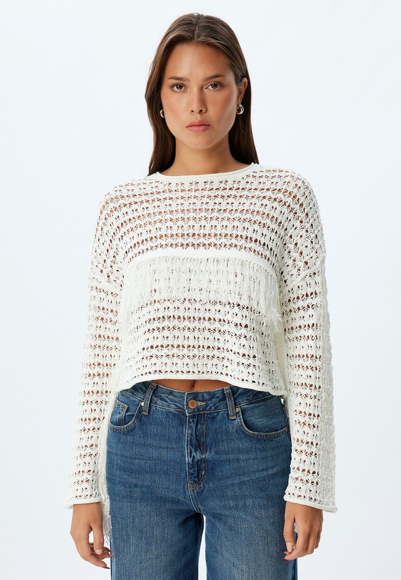 Pull en tricot blanc, court, avec un tissage ouvert texturé et des franges sur la poitrine, associé à un jean en denim bleu taille haute.