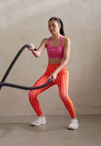 Un reggiseno sportivo rosa e leggings colorati con un motivo geometrico. La modella tiene una corda da battaglia nera in posizione di allenamento su una superficie liscia.