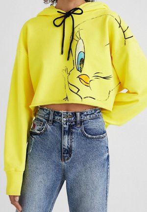 Żółta krótka bluza z kapturem z twarzą Tweety Bird i czarnymi sznurkami, zestawiona z wysokim stanem niebieskich dżinsów na stojącej osobie.