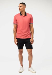 Polo rose avec des accents noirs et blancs, assorti à un short noir. Le modèle porte des baskets blanches et a des tatouages sur les bras visibles.