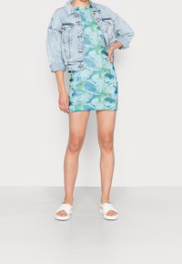 Džínová bunda s nabíranými rukávy přes mini šaty v modro-zeleným mramorovém vzoru. Bílé pantofle s širokými pásky. Hladká textura látky.