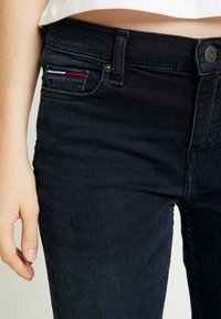 Tmavé džínové kalhoty mají přiléhavý střih s mírným texturováním. Malá kapsa má červené a bílé prošívání. Viditelné zapínání na knoflík a zip.