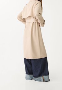 Beżowy trencz z paskiem w talii, tylnym kapturem, długimi rękawami i guzikami, w połączeniu z dżinsami o szerokich nogawkach z podwinięciem.