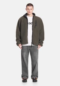 Kurtka polarowa w oliwkowej zieleni z zamkiem z przodu, dwoma bocznymi kieszeniami i małą kieszenią na piersi. Noszona na białej koszuli i szarych dżinsach.