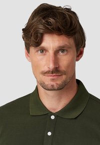 Olijfgroene polo met een klassieke kraag, een drieknops sluiting en een gestructureerde stof. Het haar is gestyled in zachte, golvende lagen.