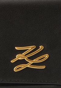 Juodas odinis piniginė su tekstūruotu paviršiumi, išsiskirianti ryškiu auksiniu logotipo emblemu priekyje. Plona, stačiakampio formos.