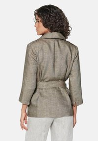 Bruine textuurjasje met een ceintuur in de taille en mouwen tot de elleboog. Met een notch-kraag en een rugsplit. G gedragen over lichtgekleurde broek.