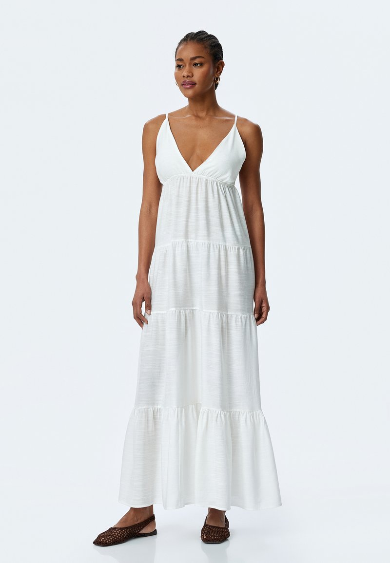 Witte maxi-jurk met een V-hals en spaghettibandjes, met een gelaagd ontwerp en een vloeiende textuur. Gecombineerd met bruine platte schoenen.