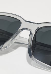 Ochelari de soare gri cu ramă din plastic transparent și lentile închise. Designul are margini netede și o formă contemporană.