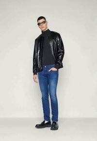 Chaqueta bomber de cuero negra con acabado texturizado, llevada sobre un suéter de cuello alto negro y jeans azules. El modelo lleva gafas de sol oscuras y zapatos negros.