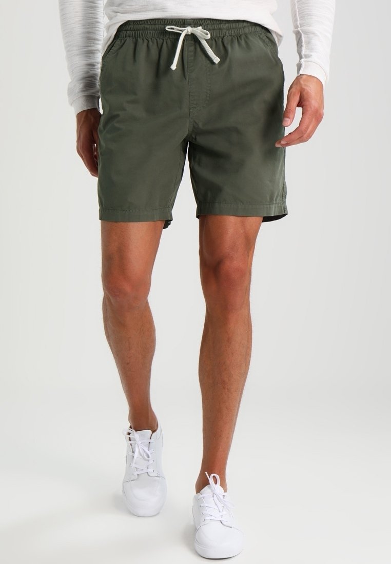 Costumi da bagno verdi con vita elastica, cordoncino, tasche laterali e vestibilità rilassata. Sneakers bianche completano il look.