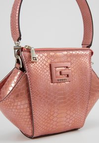 Vaaleanpunainen keinonahkainen käärmeennahkalaukku, jossa on jämäkkä muoto, kiiltävä pinta ja ylhäällä kantokahva. Varustettu näyttävällä "G"-logolla ja hopeisilla yksityiskohdilla.