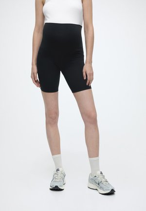 Gravid person iført sorte graviditetsbiker-shorts, hvid tanktop, hvide sokker og grå løbesko, stående mod en hvid baggrund.