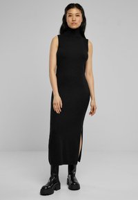Robe sans manches en tricot noir avec un col haut, une fente sur le côté et une coupe slim. Associée à des bottes compensées noires massives pour le contraste.