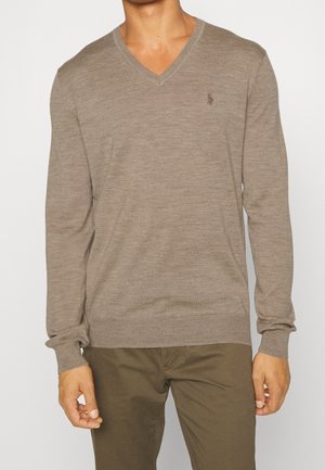 Homme portant un pull à manches longues à col en V de couleur marron clair avec un petit logo brodé sur le côté gauche de la poitrine, assorti à un pantalon olive.