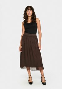 Bruine midi-rok met een vloeiende doorschijnende laag en een elastische tailleband, gecombineerd met een aansluitend zwart tanktop en zwarte hakken.