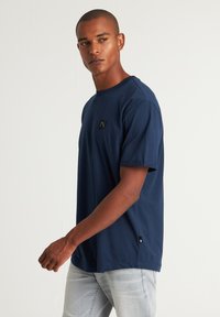 T-shirt di cotone blu navy con maniche corte, dotato di un piccolo patch logo nero e dettagli sulle cuciture laterali. Design classico a colletto rotondo.