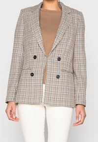 Dwurzędowa marynarka w brązowo-kremowy wzór houndstooth, z czarnymi guzikami, wyraźnie zarysowanymi ramionami oraz dwiema przednimi kieszeniami.