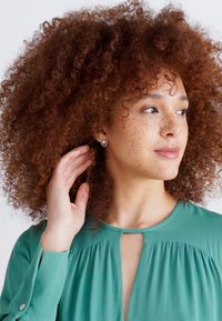 Blusa verde con scollo a chiave, tessuto morbido, maniche lunghe e vestibilità rilassata. Modella che indossa orecchini a bottone rotondi, mostrando capelli ricci.