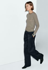 Donna che cammina con le mani in tasca, indossando una maglietta a righe con maniche lunghe, pantaloni larghi neri, scarpe nere e un bracciale in argento al polso.